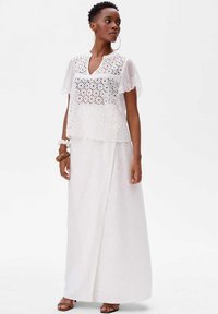 Biała haftowana bluzka z krótkimi falbankowymi rękawami, w zestawieniu z długą białą spódnicą. W bluzce wyróżniające się kwiatowe wycięcia w designie. Brązowe sandały.