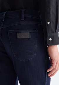 Jean Wrangler bleu foncé avec une étiquette sur la poche arrière, porté avec une chemise noire à manches longues montrant des poignets boutonnés et une main reposant sur le côté.