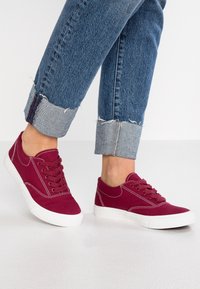 Scarpe da ginnastica in tela color bordeaux con suole in gomma bianche e occhielli. Presentano un design con lacci e cuciture a contrasto. Abbinate a jeans di denim blu arrotolati.