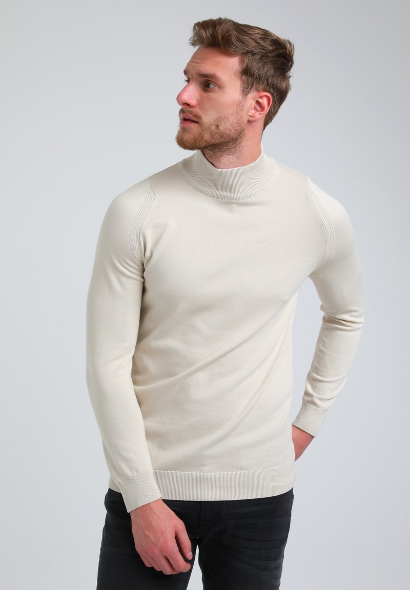 Kremna turtleneck pulover z dolgimi rokavi, rebrastim ovratnikom in manšetami, prilegajoč obliko, gladka tekstura in subtilne podrobnosti na ramenih.
