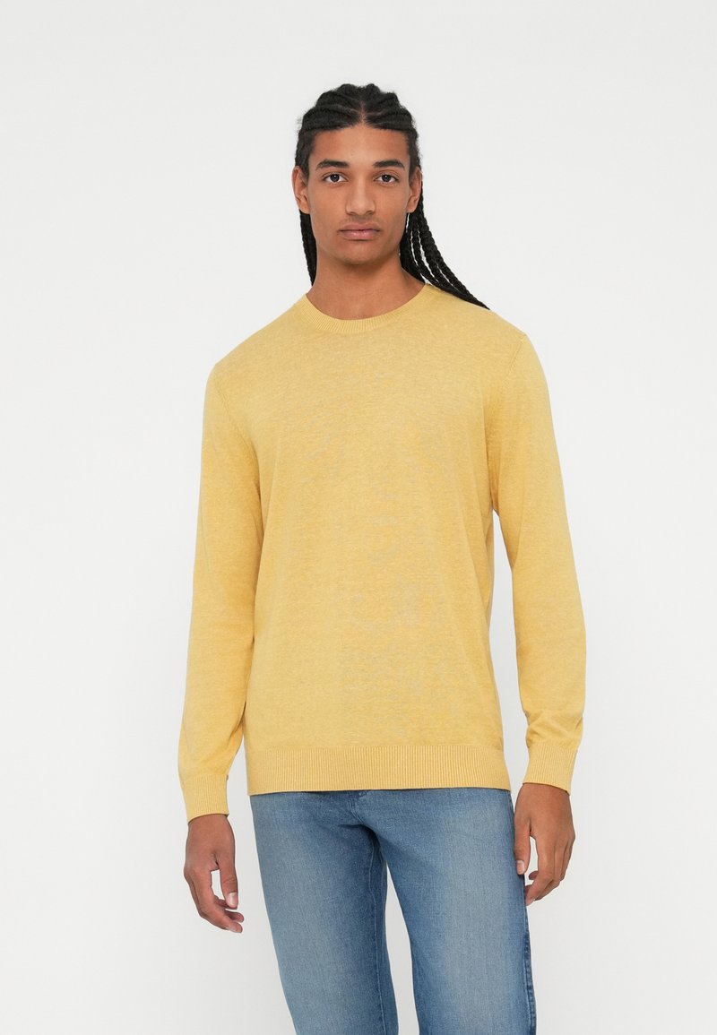Maglione giallo lavorato a maglia con maniche lunghe, scollatura rotonda e orlo a coste. Indossato con jeans azzurri chiari, caratterizzati da una texture liscia.