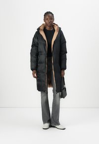 Black puffer coat with beige lining, knee-length, zip closure, and elasticated cuffs, paired with gray flared jeans and white shoes.