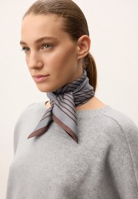 Eșarfă gri din mătase cu dungi diagonale maronii, legată la gât. Se asortează cu un pulover tricotat de un gri deschis, evidențiind textura fină și un luciu subtil.