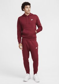 Bordeaux Nike hoodie met een voorzak en wit logo, gecombineerd met bijpassende joggerbroeken. Heeft geribbelde manchetten en enkelopeningen.