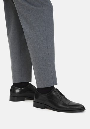 Czarne skórzane eleganckie buty o gładkim wykończeniu, zaokrąglonym nosku i wiązane na sznurówki, zestawione z jasnoszarymi, dopasowanymi spodniami.