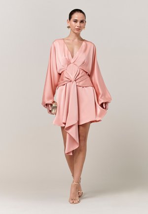 Frau trägt ein pinkfarbenes Mini-Kleid aus Satin mit langen Ärmeln, tiefem V-Ausschnitt, drapiertem Taillendetail, hält eine goldene Clutch und trägt goldene High Heels.