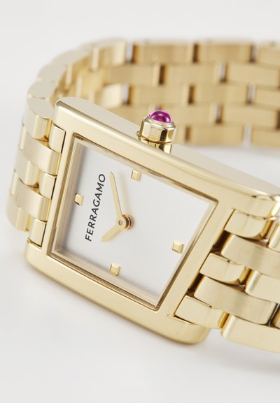 Montre rectangulaire dorée avec un cadran blanc, quatre marqueurs horaires carrés dorés et une touche de rose au niveau de la couronne. Bracelet en métal à maillons.