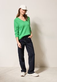 Grün, übergroßes V-Ausschnitt-Sweatshirt aus weichem Material, kombiniert mit dunkelblauen geraden Jeans. Das Model trägt eine weiße Mütze und helle Sneakers.