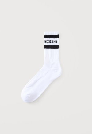 Biela členková ponožka s dvoma čiernymi vodorovnými pruhmi blízko vrchu a s čiernym slovom "MOSCHINO" medzi pruhmi.