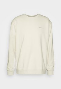 Sudadera color crema con cuello redondo, mangas largas y una textura suave. Presenta sutiles acentos de logo en el pecho y detalles en la costura lateral.