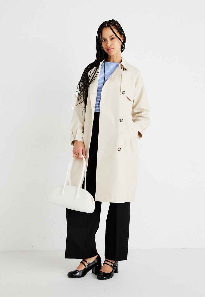 Beżowy trench z podwójnym zapięciem, w połączeniu z jasnoniebieską bluzką w paski, czarnymi spodniami o szerokich nogawkach oraz czarnymi butami na obcasie. Biała torebka.