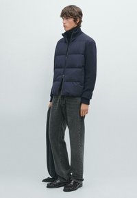 Mladý muž v námořnickém prošívaném vestě přes černý rolák, šedých džínách a černých loaferech, držící odpovídající námořnický šál.