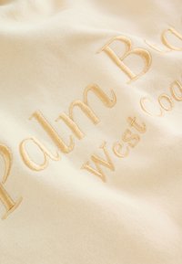Cremefarbener Stoff mit beigen, bestickten Buchstaben, die "Palm Beach West Coast" auf einer weichen, strukturierten Textiloberfläche lesen.