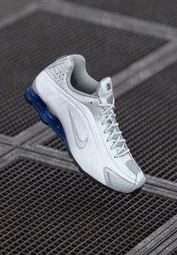 White sports shoe with light grey accents and blue rubber sole. Features lace-up design, perforated texture, and Nike logo on the side.