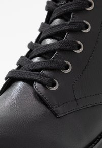 Botte en cuir noir avec lacets ronds, œillets argentés, texture lisse et détails cousus le long des bords.