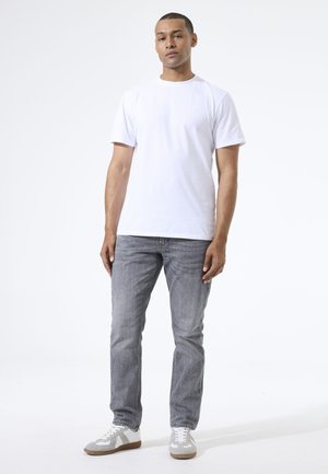Mann steht frontal zugewandt und trägt ein schlichtes weißes T-Shirt, graue Jeans und weiße-graue Sneakers vor weißem Hintergrund.