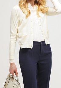 Cárdigan color crema con mangas largas, cierre de botones, dos bolsillos al frente, usado sobre una blusa blanca y pantalones azul marino. Textura suave, material ligero.