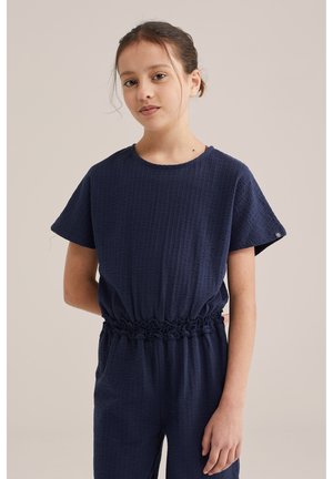 Fată tânără cu păr închis la culoare prins într-un coc jos, purtând un combinezon scurt cu mâneci dintr-un material texturat, culoarea albastru închis, cu talie strânsă, pe un fundal simplu.