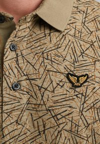 Beige polo tričko se strukturovaným vzorem černých a hnědých linií, se černým a zlatým vyšitým logem na hrudi a černými knoflíky.