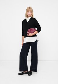 Mujer rubia con un suéter negro de pico, camisa blanca, pantalones de pierna ancha oscuros, zapatos negros, sosteniendo un bolso burdeos contra un fondo blanco.