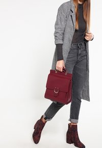 Bolso burdeos con asa superior y cierre, abrigo de cuadros grises, camiseta negra y jeans oscuros; botas de tobillo con un tacón ancho y estampado.