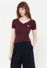 Donna con capelli rossi che indossa una maglietta attillata bordeaux a collo a V con un logo a triangolo rosso e pantaloni neri larghi, in piedi con una mano in tasca.
