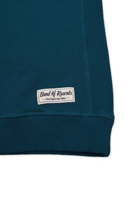 Teal ribbet sweatshirt-kant med et hvidt rektangulært mærke, hvor der står "Band of Rascals, Finest Organic Kids Clothes" med sort skrift.