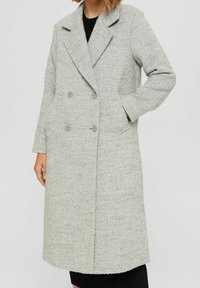 Long light gray textured double-breasted coat with notch lapels, front pockets, and four buttons, worn over a black ribbed garment.