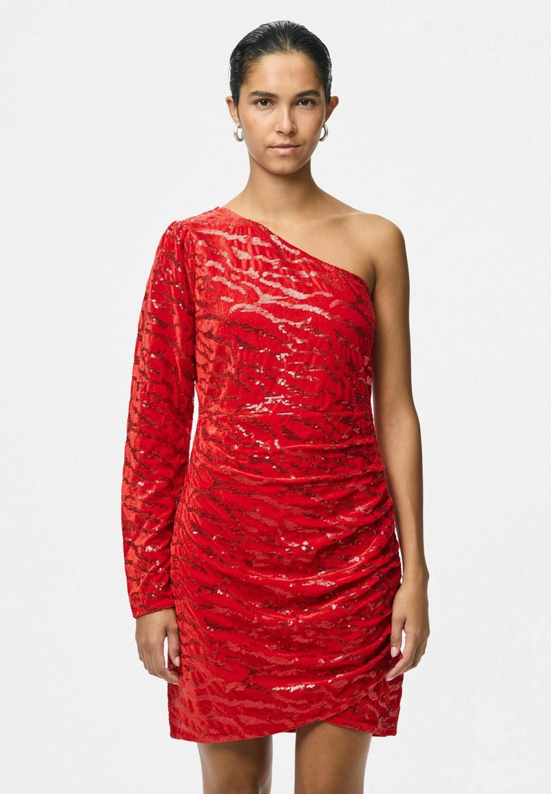 Raudona suknelė su vienu petimi, dekoruota blizgučiais, raukinėto audinio ir asimetrinio dizaino. Viena pusė turi ilgą rankovę, o siluetas yra prigludęs.