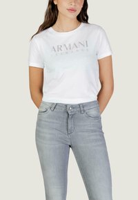 Femme portant un t-shirt blanc Armani Exchange et un jean gris clair, debout avec les mains derrière le dos sur un fond uni.