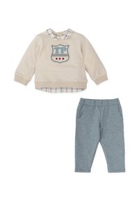 Sudadera beige con parche "TP" y estrellas rojas, superpuesta sobre un cuello a cuadros azul y blanco. Pantalones de chándal grises con dos bolsillos laterales.