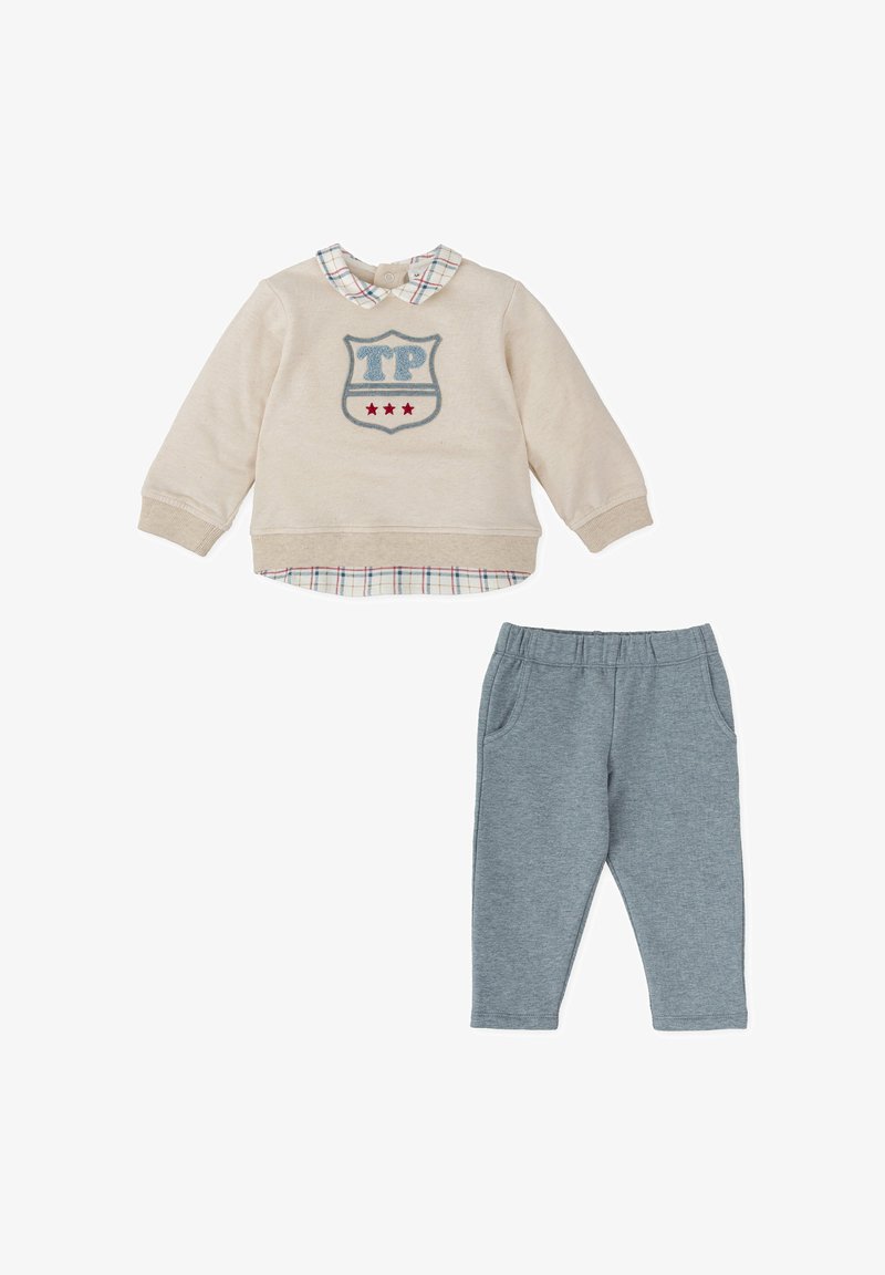 Sudadera beige con parche "TP" y estrellas rojas, superpuesta sobre un cuello a cuadros azul y blanco. Pantalones de chándal grises con dos bolsillos laterales.