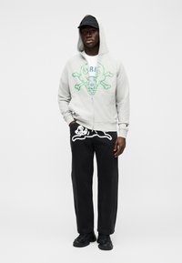 Ανοιχτό γκρι φούτερ με φερμουάρ και πράσινο γραφικό σχέδιο· λευκό μπλουζάκι από μέσα· μαύρο παντελόνι με λευκό περίγραμμα σχεδίου· μαύρο καπέλο και αθλητικά παπούτσια.