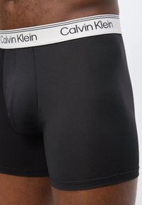 Μαύρα ανδρικά εσώρουχα boxer με λευκή ελαστική μέση που φέρει το λογότυπο Calvin Klein, φορεμένα από άτομο με σκούρο δέρμα.