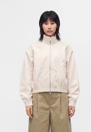 Donna con capelli neri lisci che indossa una giacca beige chiaro con zip e pantaloni larghi color kaki, in piedi davanti a uno sfondo bianco semplice.