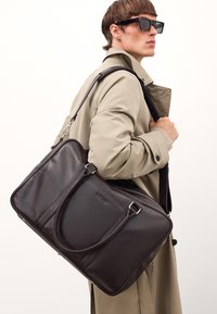 Mørkebrun lædertaske med tekstureret overflade, dobbelte håndtag, aftagelig skulderrem og subtilt logodesign. Klassisk rektangulær form.