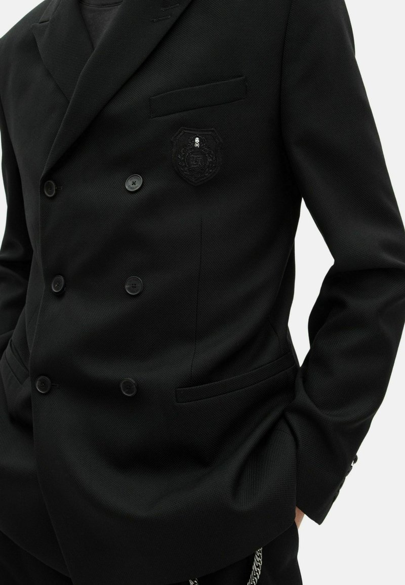 Zwarte dubbel-gebreide blazer met een gestructureerde stof, voorzien van een enkele borstzak, zwarte knopen en een gegraveerd embleem aan de linkerkant.