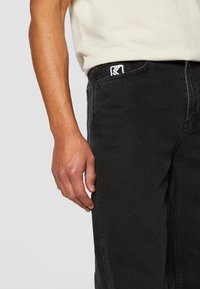 Black denim pants with a relaxed fit, featuring a subtle texture, visible stitching, and a logo patch on the waistband.