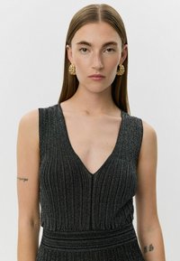 Zwarte, geribbelde jurk met een diepe V-halslijn en een mouwloos design. Heeft een aansluitende taille en een subtiele glans. Gouden ringoorbellen accentueren de look.