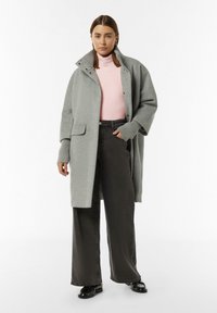 Grau Wolle Mantel mit großem Kragen, Druckknöpfen und aufgesetzten Taschen, getragen über einem pinken Rollkragenpullover und grauen weiten Jeans. Schwarze Schuhe.