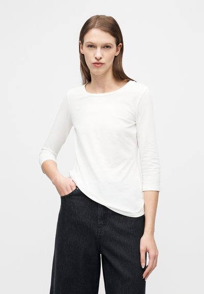 Γυναίκα με ίσια καστανά μαλλιά, φορώντας λευκό πουκάμισο με μανίκια μέχρι τον αγκώνα και μαύρο παντελόνι με φαρδιά μπατζάκια, στέκεται με το ένα χέρι στην τσέπη.