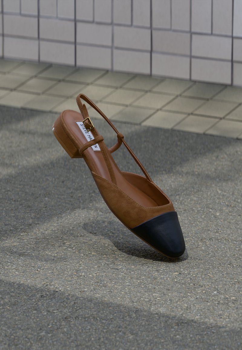 Barna lapos slingback cipő fekete hegyes orral és arany csattal, texturált járda felett lebegve, csempézett fal közelében.