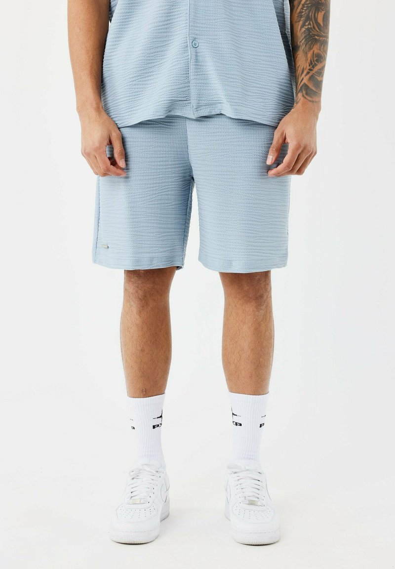 Pantalones cortos casuales de color azul claro y camisa de manga corta a juego, hechos de un tejido texturizado, similar al waffle. Llevados con zapatillas blancas y calcetines hasta la pantorrilla.
