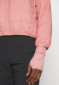 Veste courte matelassée rose avec fermeture éclair à l'avant, texture quiltée et poignets élastiques, associée à un pantalon noir. Dispose d'un trou pour le pouce dans la manche.