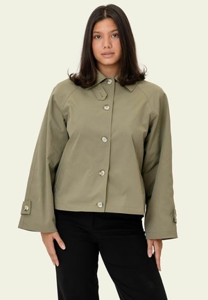 Mladá žena s rovnými tmavými vlasmi, oblečená v olivovo zelenej zapínacej bunde a čiernych nohaviciach, stojaca na neutrálom pozadí.