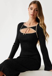 Abito nero a costine con maniche lunghe, caratterizzato da un colletto con apertura impreziosito da un fiocco beige. Tessuto liscio con una veste aderente.