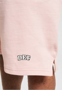 Shorts en coton rose avec un logo brodé "DEF" en noir. Les détails notables comprennent des fentes latérales et une finition douce et texturée.