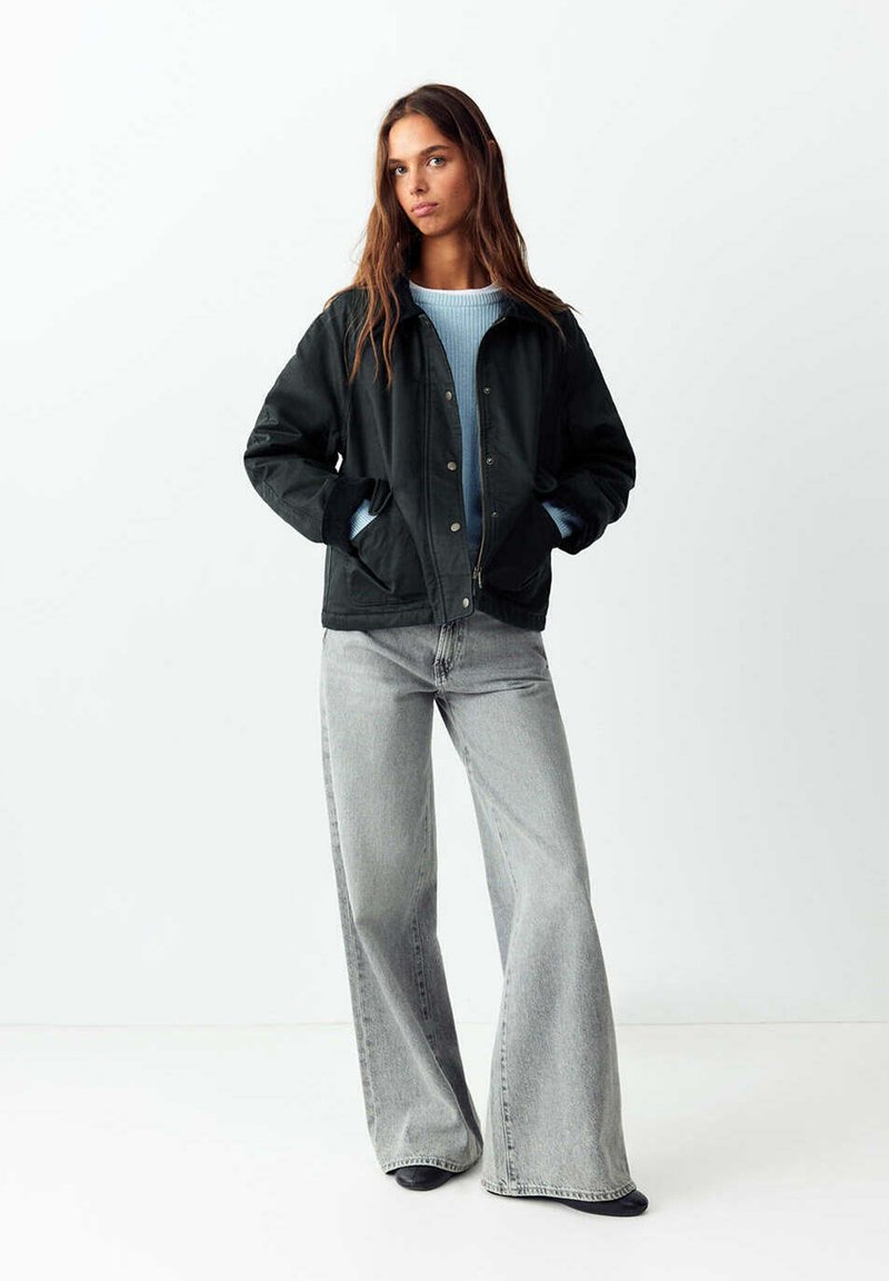 Jeune femme debout, les mains dans les poches, portant une veste noire, un pull bleu clair et un jean gris à jambes larges sur un fond uni.