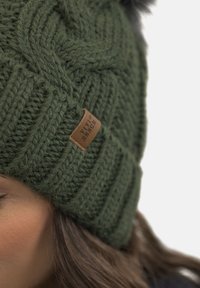 Gorro de punto verde con un patrón texturizado, que presenta una etiqueta de cuero en el lado. La parte superior tiene un pompón negro como acento.