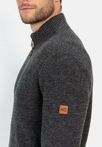 Dunkelgrauer gestrickter Pullover mit hohem Kragen, ausgestattet mit einem Reißverschluss und einem Logo-Patch aus hellbraunem Leder am Ärmel. Strukturierter Stoff mit gerippten Details.